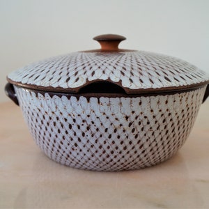May include: A white ceramic serving bowl with a lid. The bowl has a brown rim and handles, and a lattice pattern. The lid has a small brown knob. The bowl is likely used for serving food.