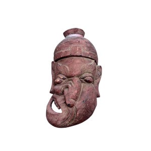 Puede incluir: Escultura de madera de color marrón rojizo desgastado que representa la cabeza de un hombre con el rostro severo. La talla presenta un sombrero detallado, cejas fruncidas y una expresión distintiva con la boca abierta. Fondo blanco.