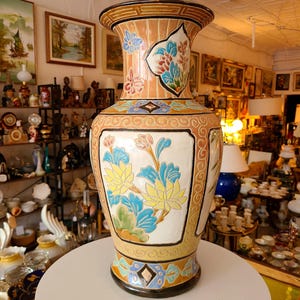 Może przedstawiać: Duży, ozdobny wazon ceramiczny z kwiatowym wzorem. Wazon jest pomalowany w odcieniach niebieskiego, żółtego, zielonego i różowego. Ma brązową i złotą podstawę oraz szeroki otwór.