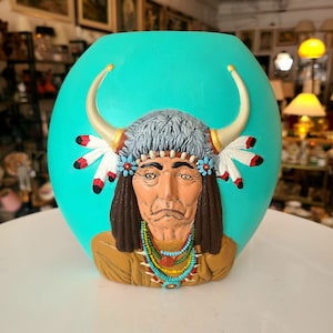 May include: A turquoise ceramic vase featuring a relief sculpture of a Native American man wearing a headdress with feathers and beads. The man has a serious expression and is looking straight ahead.
