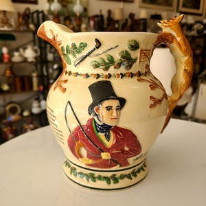 Peut inclure: Une cruche en céramique avec une poignée en forme de renard. La cruche est décorée d'une scène de chasse représentant un homme en veste rouge et un chapeau haut de forme noir. La scène comprend un chien de chasse, un fouet et une pipe.