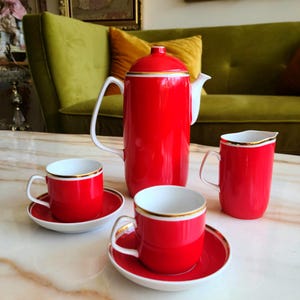 May include: A red and white porcelain tea set with a teapot, creamer, and two cups and saucers. The set has a gold trim around the edges.
