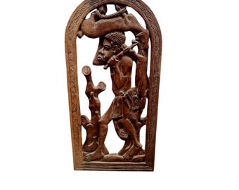 May include: A carved wooden relief sculpture showing a figure playing a flute, with an animal above. The sculpture is framed with an arched top and intricate border details. The wood has a warm brown tone.