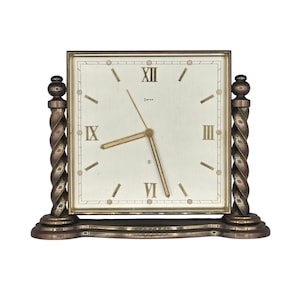 May include: A square table clock with a cream-colored face, Roman numerals, and the word "SWIZA" printed below the twelve o'clock position. The clock is framed by two bronze-toned, twisted spiral columns on a matching base with gold-toned hands.