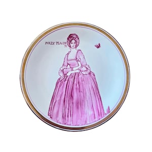 May include: A decorative plate with a pink illustration of a woman in a historical gown, with the text "POLLY PEACHY" and a gold rim. A pink butterfly is also depicted on the plate.