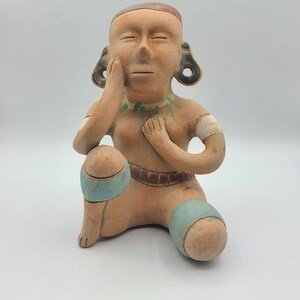 May include: A terracotta figurine of a person sitting with their legs crossed, wearing a blue and brown patterned skirt and a green necklace. The figurine has a brown face with closed eyes and a hand resting on their chin.