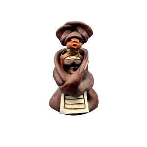 Op de afbeelding: Handgemaakte keramische figuur van een vrouw, met een donkerbruine hoofddoek en sjaal. De figuur heeft een bruin gezicht, een goudkleurige halsring en een wit en bruin schortachtig detail. Op een witte achtergrond.