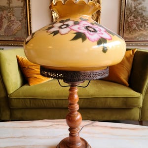 May include: A vintage table lamp with a yellow glass shade featuring pink and white floral designs. The lamp has a wooden base with a decorative metal trim.