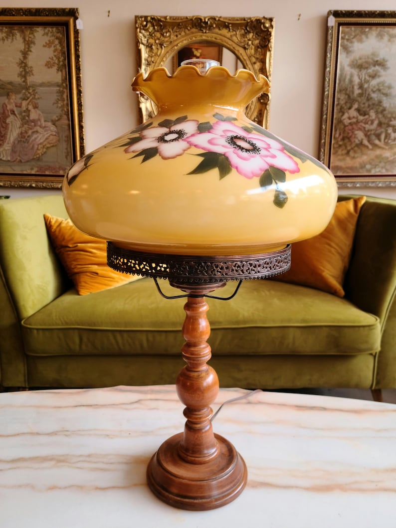 May include: A vintage table lamp with a yellow glass shade featuring pink and white floral designs. The lamp has a wooden base with a decorative metal trim.