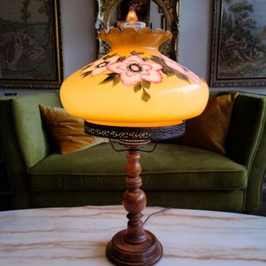 May include: A vintage table lamp with a yellow glass shade featuring hand-painted pink flowers. The lamp has a wooden base with a decorative metal trim around the shade.