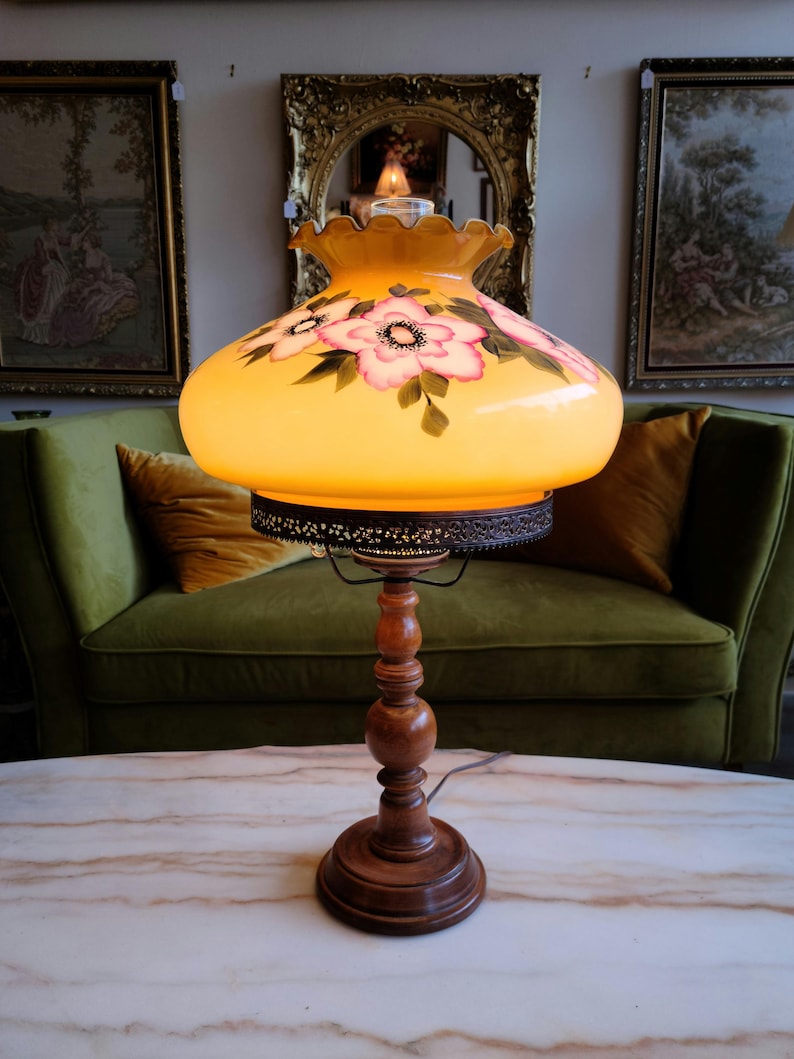 May include: A vintage table lamp with a yellow glass shade featuring hand-painted pink flowers. The lamp has a wooden base with a decorative metal trim around the shade.