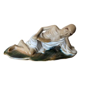 May include: A ceramic figurine of a reclining figure with a bald head, tan skin, and closed eyes, resting on one elbow. The figure is draped in a white cloth and holds a small tan object, positioned atop a dark green, leaf-shaped base.