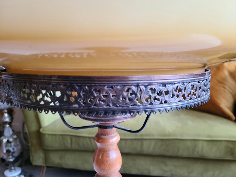 May include: A vintage table lamp with a round, scalloped, amber glass shade. The shade has a decorative metal trim with an intricate floral design. The lamp has a wooden base with a turned pedestal.
