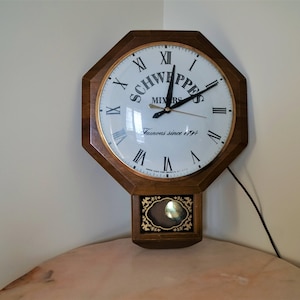 Puede incluir: Un reloj de pared octogonal de madera con una esfera blanca y marcadores de hora romanos negros. El reloj tiene un péndulo negro y un logotipo dorado de Schweppes Mixers con el texto "Famous since 1791".