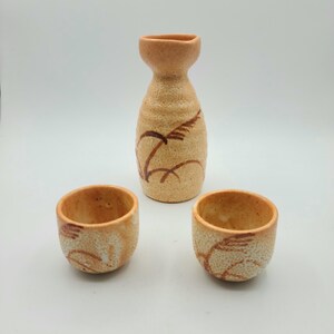 Puede incluir: Un juego de tres tazas de sake de cerámica y una botella de sake. Las tazas y la botella están decoradas con un diseño de hierba marrón y blanco.