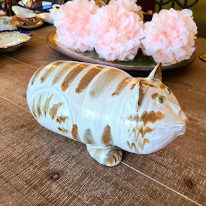 Peut inclure: Une figurine de chat en céramique avec des rayures brunes et blanches. Le chat a une grosse tête et une large bouche ouverte. La figurine est glacée d'un glacis blanc et brun.