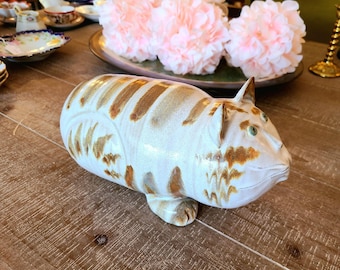 May include: A ceramic cat figurine with brown and white stripes. The cat has a large head and a wide, open mouth. The figurine is glazed in a white and brown glaze.