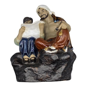 May include: A glazed ceramic figurine depicting two figures sitting on a dark, textured rock base. One figure wears a white head covering and brown trousers, while the other wears dark blue trousers and holds a white, open document or map.