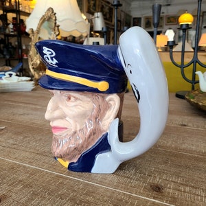 May include: A blue and white ceramic mug shaped like a sailor with a white whale tail handle. The sailor has a yellow hat with a gold anchor emblem and a brown beard.