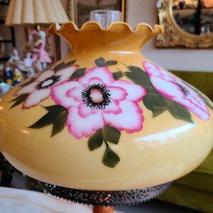 May include: A yellow glass lampshade with a ruffled edge and painted pink and white flowers. The lampshade has a black metal base.