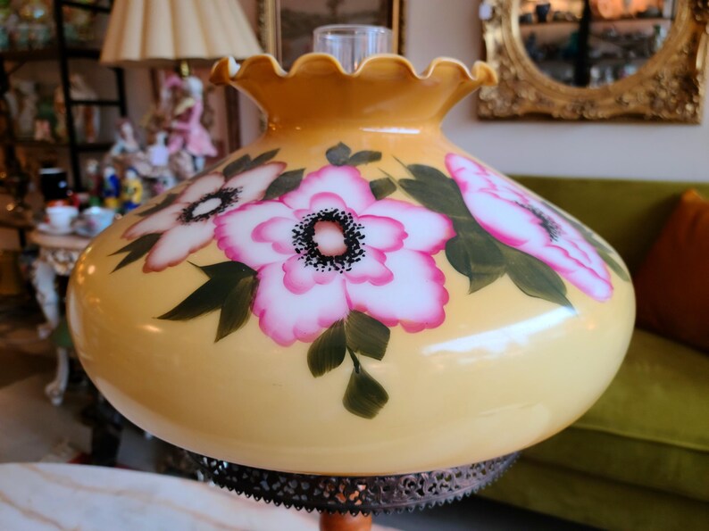 May include: A yellow glass lampshade with a ruffled edge and painted pink and white flowers. The lampshade has a black metal base.