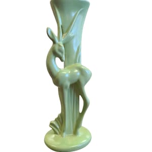 May include: A light green ceramic vase featuring a deer figurine. The vase has a tall, slender form with a deer sculpted on the side. The deer stands beside a vertical element. The vase has a round base and is approximately 25 cm tall.