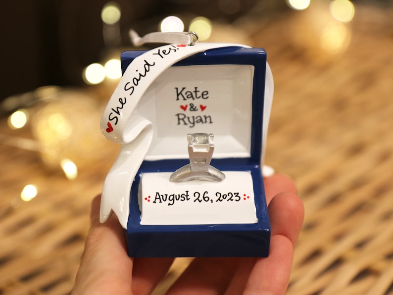 Op de afbeelding: Een blauwe ringdoos ornament met een wit lint waarop "She Said Yes!" staat. De doos is open en bevat een zilveren ring. De doos is gepersonaliseerd met de namen "Kate & Ryan" en de datum "August 26, 2023".