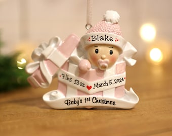 May include: Pink and white personalized baby's first Christmas ornament. The ornament features a baby girl in a pink hat and a pacifier, peeking out of a pink and white gift box. The ornament is decorated with the baby's name, birthdate, and weight: "Blake", "March 5, 2024", and "7 lbs 13 oz".