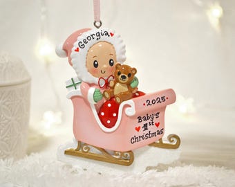 May include: A pink Christmas ornament featuring a baby in a Santa hat and a teddy bear in a sleigh. The ornament has the text "Georgia" and "Baby's 1st Christmas" with the year "2025" on it.