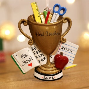 May include: A gold-colored trophy ornament with the words "Best Teacher." It features a ruler, pencils, scissors, and a small book with the name "Janet" and a heart. A red apple and a pencil are also included. The base reads "2021."