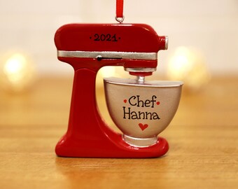 May include: Red and silver stand mixer Christmas ornament with a silver bowl that says "Chef Hanna" with a red heart. The mixer has a black label that says ".2021."