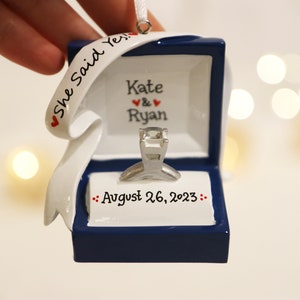 Op de afbeelding: Een blauwe en witte keramische ornament in de vorm van een ringdoos met een zilveren ring erin. De ornament heeft een wit lint met de tekst "She Said Yes." en de namen "Kate & Ryan" en de datum "August 26, 2023".