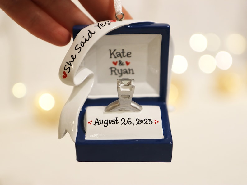 Op de afbeelding: Een blauwe en witte keramische ornament in de vorm van een ringdoos met een zilveren ring erin. De ornament heeft een wit lint met de tekst "She Said Yes." en de namen "Kate & Ryan" en de datum "August 26, 2023".