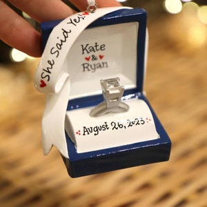 Op de afbeelding: Een blauwe ringdoos-versiering met een witte ring en een wit lint met de tekst "She Said Yes!" met rode hartjes. De doos heeft ook de namen "Kate & Ryan" en de datum "August 26, 2023" erop.