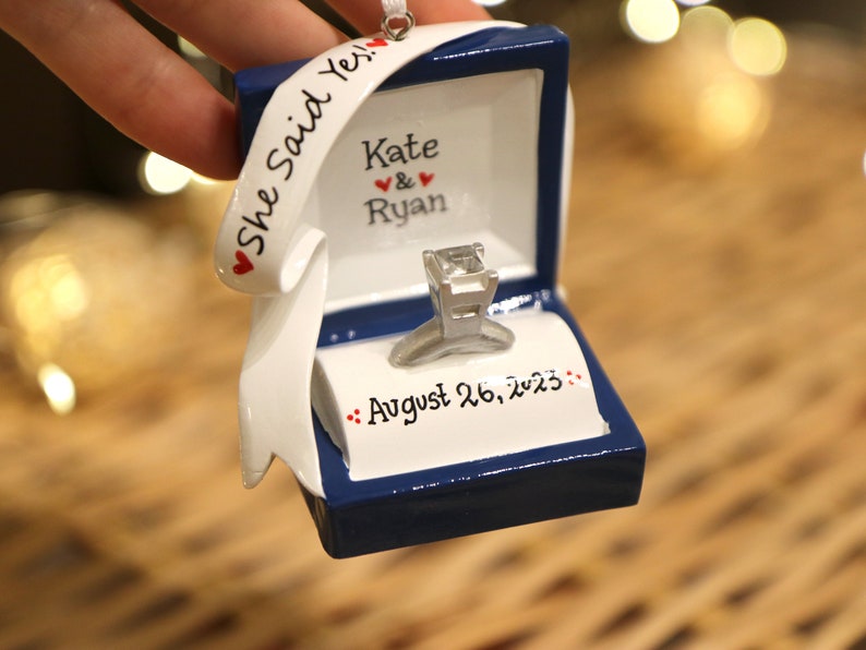 Op de afbeelding: Een blauwe ringdoos-versiering met een witte ring en een wit lint met de tekst "She Said Yes!" met rode hartjes. De doos heeft ook de namen "Kate & Ryan" en de datum "August 26, 2023" erop.