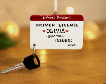 Può includere: Ciondolo bianco e rosso a forma di patente di guida con il testo "Drivers Beware! Driver License Olivia New York Issued: 2021" e una piccola chiave nera attaccata.