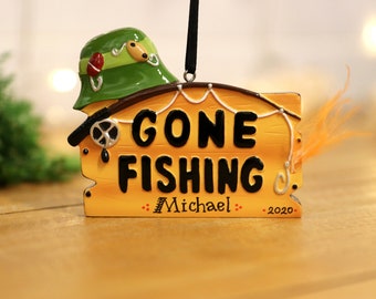 Könnte beinhalten: Ein gelbes Holzschild mit dem Text "Gone Fishing" in schwarzen Buchstaben. Das Schild hat einen grünen Fischerhut, eine Angelrute und einen Angelköder mit orangefarbenen Federn. Das Schild hat auch den Text "Michael" und "2020" in schwarzen Buchstaben.
