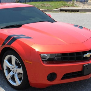 Puede incluir: Un Chevrolet Camaro rojo con rayas de carreras negras en el capó y los guardabarros. El coche tiene una parrilla negra y una placa de matrícula negra.