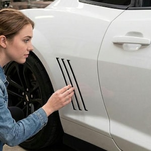 May include: A white sports car with black accents, including black wheels and a series of black vertical vents on the side. The car's door handle is visible, and the vehicle appears to be in a garage or similar setting.