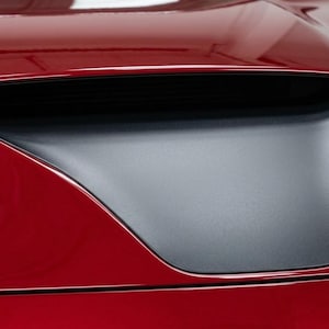 May include: Close-up of a red car hood with a black air intake. The hood has a glossy finish and a sleek, aerodynamic design. The air intake is a matte black color and is integrated into the hood's design.