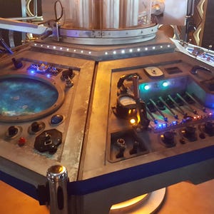 May include: A silver control panel with a blue and silver rim. The panel has a variety of buttons, knobs, and lights. The panel is lit with blue and white lights.