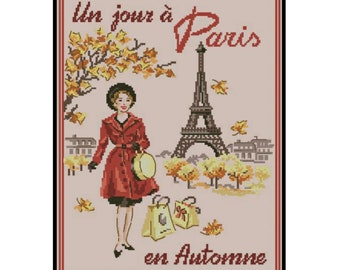 May include: Cross-stitch design featuring a woman in a red coat, holding a hat, with the Eiffel Tower in the background. The text reads "Un jour à Paris en Automne". Autumn leaves and shopping bags complete the scene.