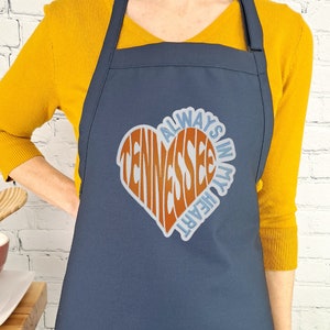 May include: A navy blue apron with a large heart-shaped design that says "Always in my heart Tennessee".