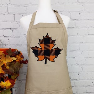 May include: Tan apron with a large leaf design. The leaf is outlined in orange and filled with a black and orange plaid pattern. The apron has a neck strap and two front pockets. The background is a white brick wall.