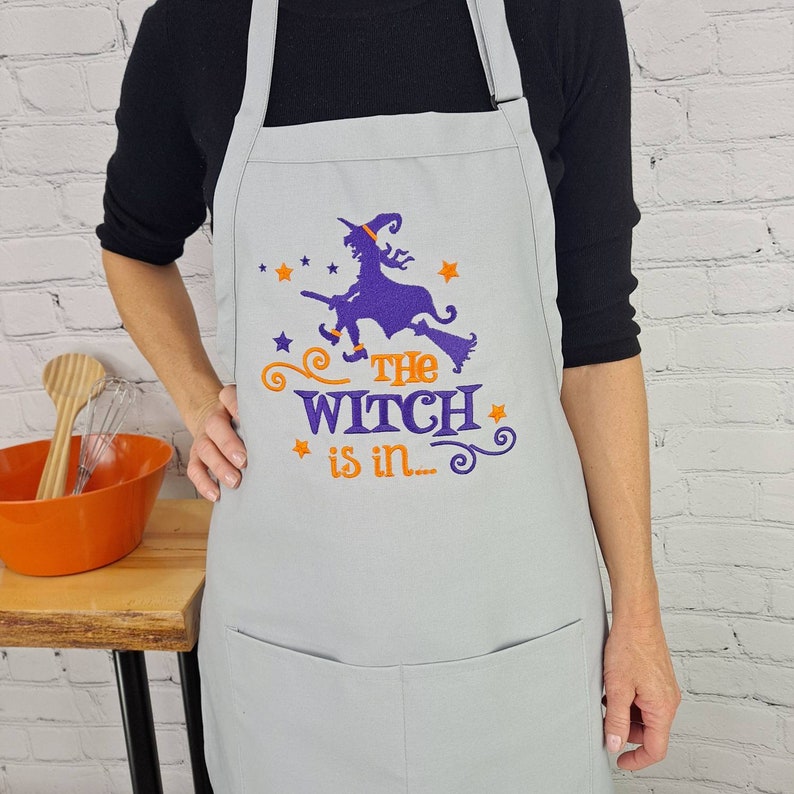 Puede incluir: Un delantal de cocina gris con un dise&ntilde;o bordado en morado y naranja de una bruja volando en una escoba con el texto "The Witch is in..."