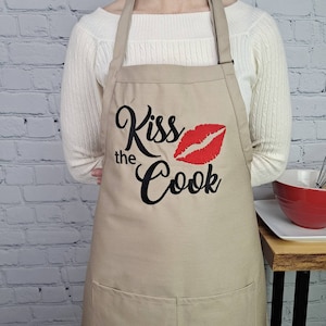 May include: Beige kitchen apron with a black embroidered design that says "Kiss the Cook" with a red embroidered lip print.