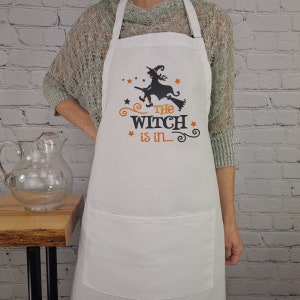 Puede incluir: Delantal de cocina blanco con una silueta negra de una bruja volando en una escoba. El texto "The Witch is in..." est&aacute; impreso en naranja en el delantal.