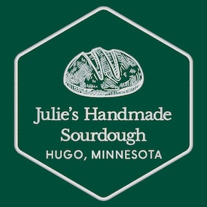 May include: A white hexagonal logo with a white outline on a dark green background. The logo features a hand-drawn illustration of a loaf of sourdough bread and the text "Julie's Handmade Sourdough Hugo, Minnesota".