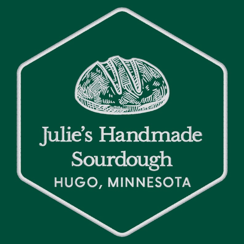 May include: A white hexagonal logo with a white outline on a dark green background. The logo features a hand-drawn illustration of a loaf of sourdough bread and the text "Julie's Handmade Sourdough Hugo, Minnesota".