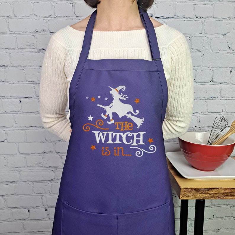Puede incluir: Delantal de cocina morado con una silueta blanca de una bruja volando en una escoba. El texto "The Witch is in..." est&aacute; impreso en naranja debajo de la bruja.